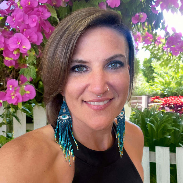 Headshot of Breanna with blue earrings in front of pink flowers and a white picket fence. She is a spiritual life coach, day maker, psychic and healer