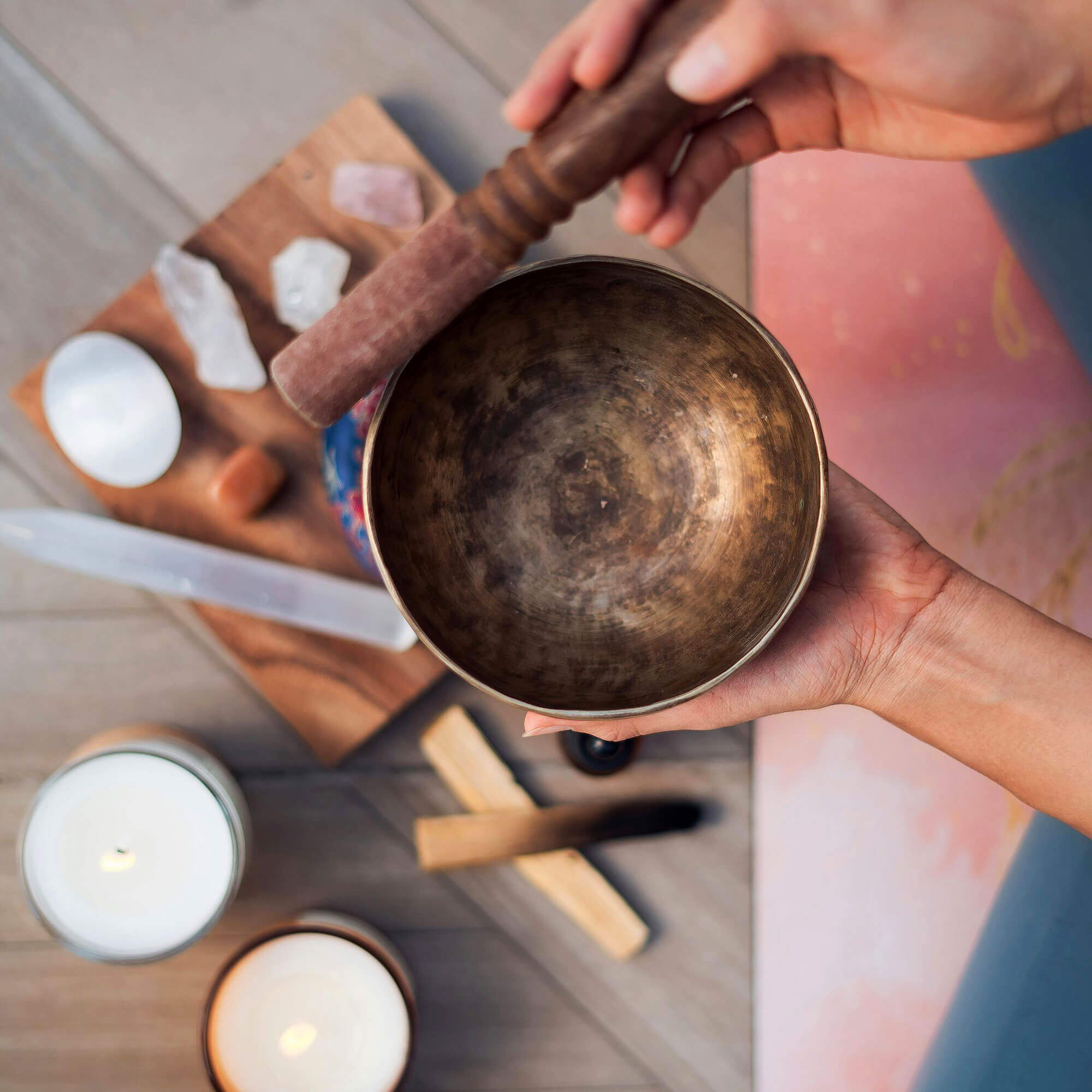 Person holding a bronze singing bowl with a wooden mallet, surrounded by candles and crystals on a wooden surface.